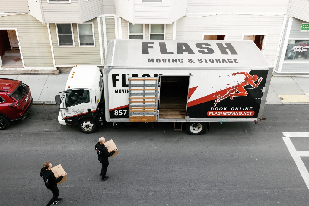 Two professional movers carrying boxes into a Flash Moving & Storage truck on a residential street in Boston. Reliable college student movers providing stress-free dorm and off-campus apartment moving services