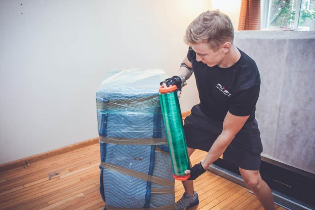 Professional mover from Flash Moving & Storage wrapping a piece of furniture with protective padding and plastic wrap in a Boston apartment. Expert college student moving services ensuring safe and secure relocation